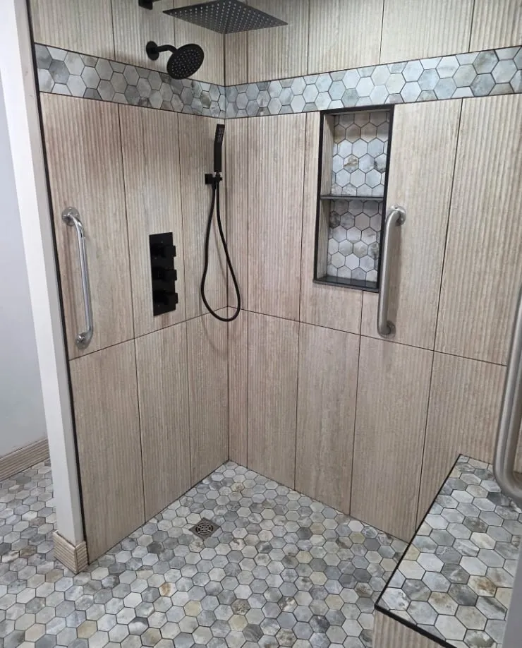 A functional walk-in shower built for a disabled veteran — wood-look tile, a hex stone accent band, grab bars, niches and a corner bench.