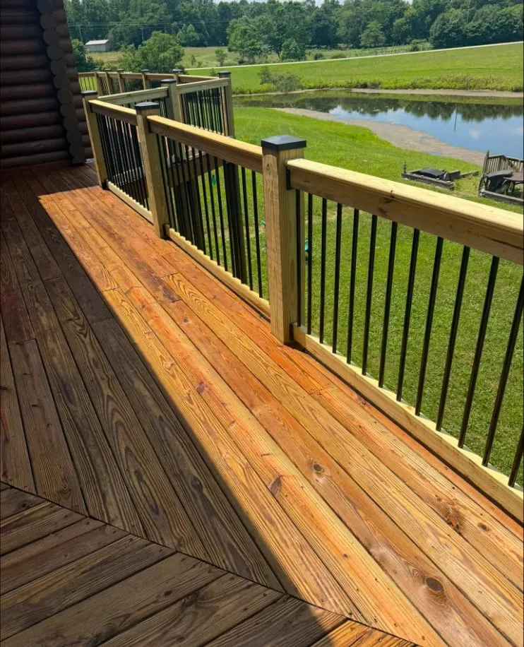 Pressure-treated decking with cedar-tone rails and black balusters, wrapping a log home above its pond.